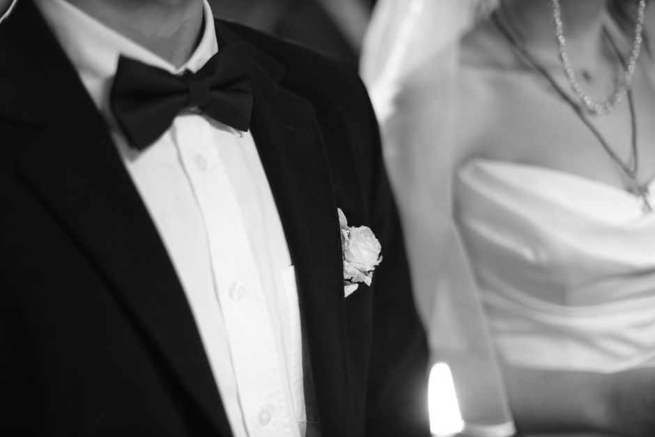 Close-up of a man's torso dressed in a classic black tuxedo with a white shirt and black bow tie, featuring a white floral boutonniere pinned to the lapel. Next to him, a woman wearing a strapless whi