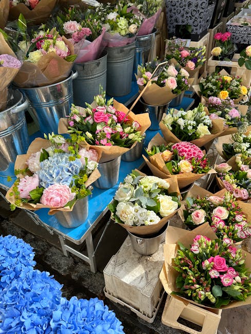 A display of fresh flower bouquets arranged in metal buckets and wrapped in brown paper, featuring a variety of pastel pink roses, white and pale green foliage, light blue hydrangeas, and other mixed