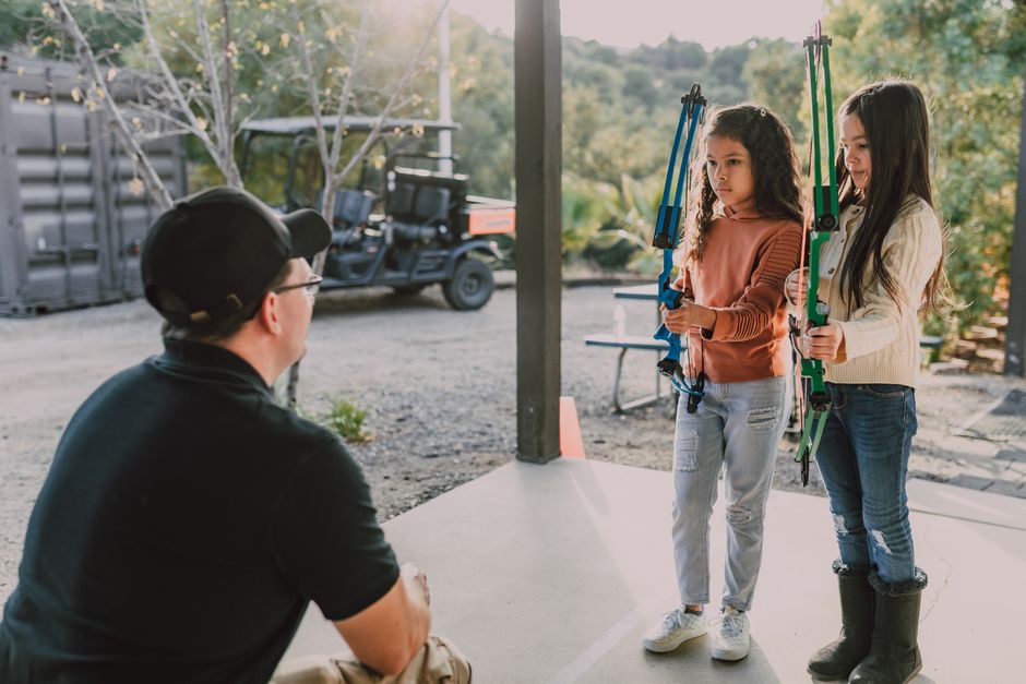 Two young girls with long dark hair, one wearing a beige sweater and jeans, the other in a rust-colored top and light blue jeans, are standing outdoors under a shelter holding colorful archery bows an