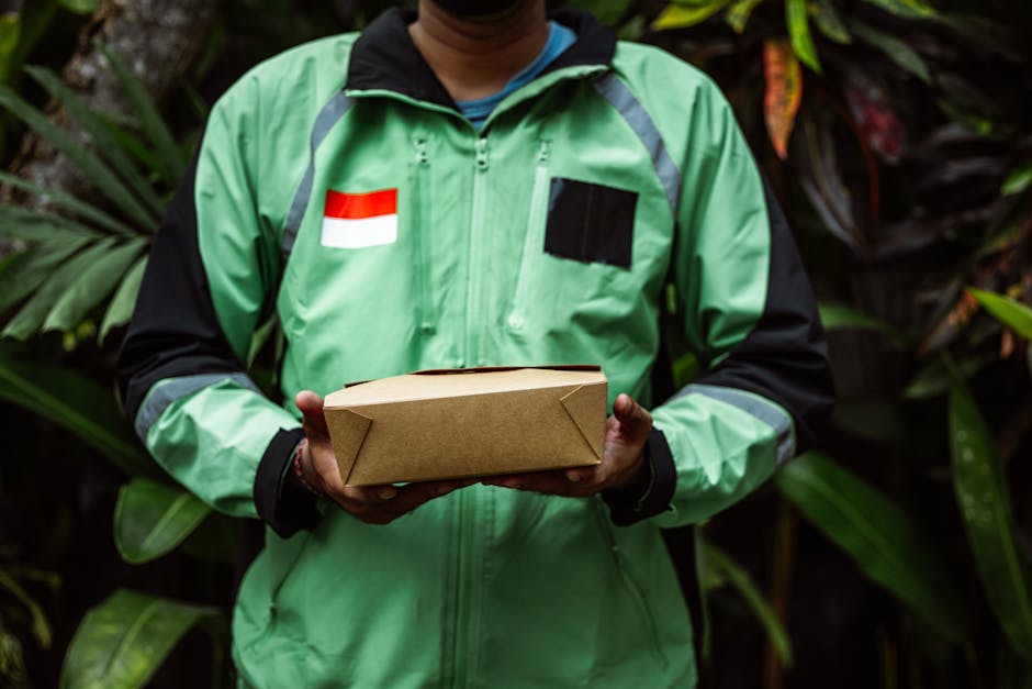A delivery person wearing a green jacket with black and grey accents, standing outdoors surrounded by lush green foliage. The individual is holding a neatly wrapped, rectangular cardboard parcel, like