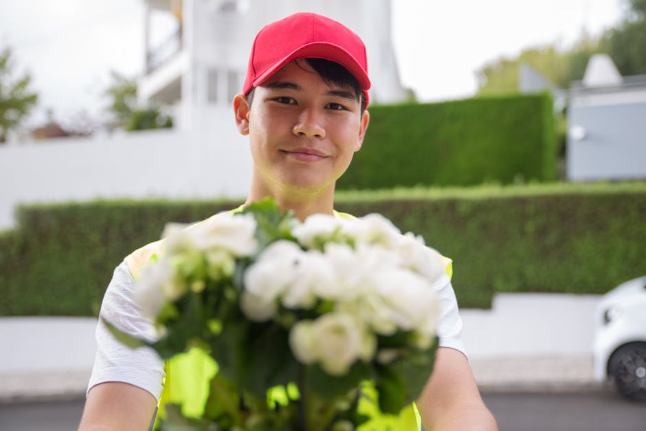 A delivery person wearing a red cap and bright yellow high-visibility vest is holding a bouquet of fresh white flowers, including roses and lilies, wrapped in clear plastic with a gold ribbon. The bou
