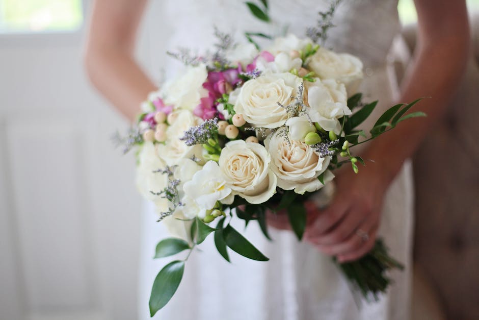 A person holding a floral bouquet featuring large white roses, delicate pale pink roses, small white ranunculus, lavender sprigs, and light purple accent flowers, all arranged with greenery. The bouqu