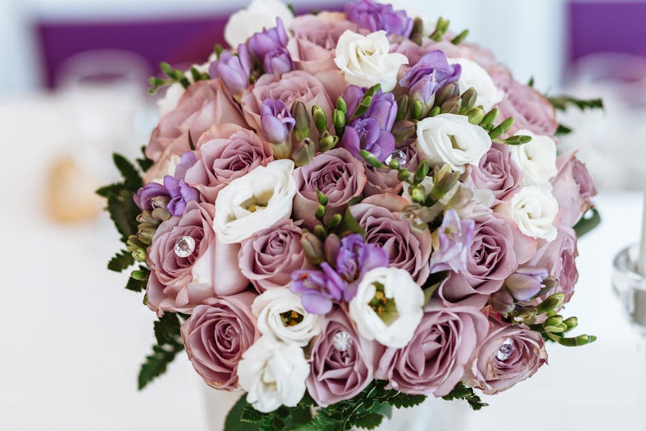 A close-up image of a fresh floral arrangement featuring a round bouquet of pastel pink roses, white eustoma, and purple lisianthus, accented with small green leaves and flower buds. The bouquet is wr
