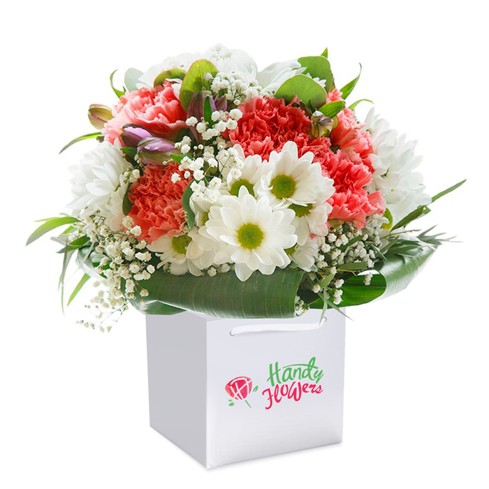 A captivating bouquet brimming with life and colour is presented within a clean, square white gift bag, showcased against a stark white background that enhances every detail. This arrangement, expertly crafted by Bow's experienced florists, features a rhapsody of soft coral and peach carnations whose ruffled, plush petals ripple outwards, mingling with spirited scarlet accents. Sprays of crisp white daisies with vivacious yellow-lime centers inject sunlight and cheer, reminiscent of afternoons in Mile End Park, while wisps of delicate baby's breath are scattered throughout to add gentle, cloud-like texture. Peeking from the bouquet's left side are several purple-tipped flower buds, subtle notes of promise and fresh beginnings. Encircling the blooms is a ring of abundant, dark green foliage, broad and glossy, forming an elegant base reminiscent of the greenways tracing the Lea River. The gift bag's soft ribbon handles arch gracefully overhead, suggesting a bouquet ready for delivery-perhaps hand-delivered around Old Ford or through the historic streets near Bow Church. This artfully arranged composition is not only visually striking, but also exudes the sense of newness, abundance, and sophistication essential for birthdays, anniversaries, or simply sharing a heartfelt sentiment with someone dear. With its intricate balance of colour, freshness, and carefully arranged florals, this bouquet reflects the insight and community spirit unique to Bow's local flower shops.